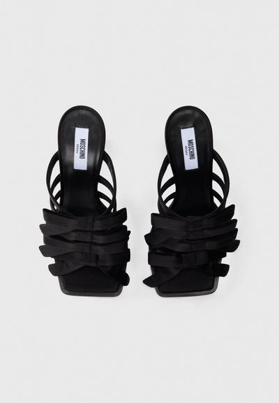 Sandales à enfiler en cuir noir Moschino avec bout carré, brides en nœud superposées et fines sangles arrière sur fond blanc.