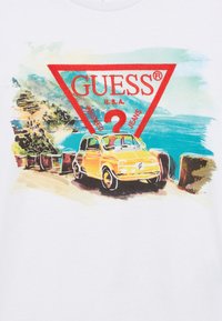 Λευκό μπλουζάκι με πολύχρωμο γραφικό τύπωμα που απεικονίζει ένα κίτρινο βίντατζ αυτοκίνητο και το λογότυπο "GUESS®" μέσα σε κόκκινο τρίγωνο, με φόντο παραθαλάσσιο τοπίο.