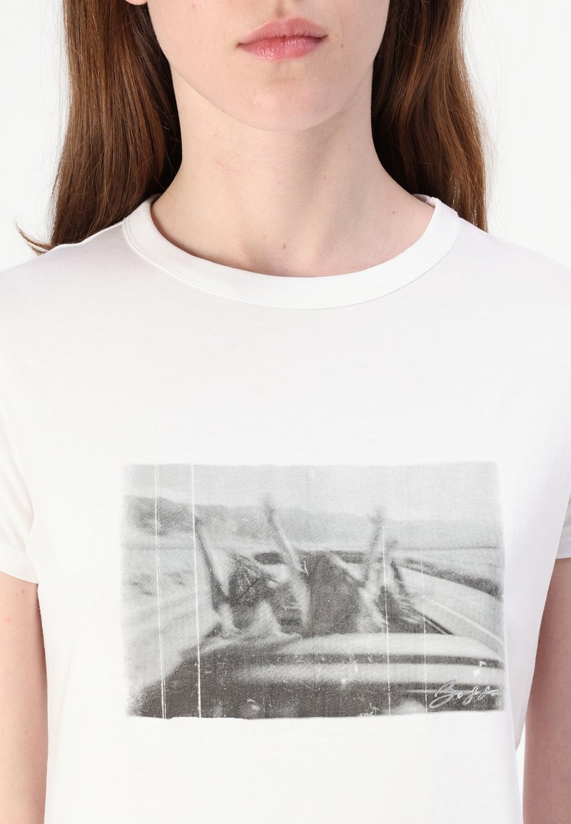 Weißes Baumwoll-T-Shirt mit einem schwarz-weißen Grafikdruck von zwei Figuren mit erhobenen Armen, am unteren Rand mit einer Unterschrift versehen.