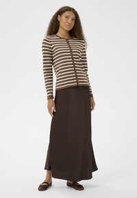 Kvinna med lockigt hår iklädd en brun och beige randig knappad cardigan, lång mörkbrun kjol och bruna lägenheter med svarta remmar.