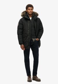 Parka negra aislada con capucha forrada de piel, cierres de cremallera y a presión, dos bolsillos frontales, superpuesta sobre un suéter gris y una camisa de cuadros. Jeans oscuros y zapatos marrones.