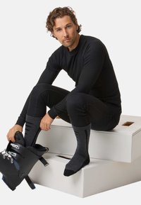 Schwarzes thermisches Basisschicht-Oberteil und Hose mit geripptem Muster, graue Skisocken und schwarze Skistiefel werden getragen. Sitzt auf einer weißen Plattform.