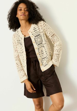 Moteris su garbanotais plaukais, vilkinti kreminės spalvos megztą su sagomis perliemenuotą megztinį ant tamsaus tankinio ir laisvų juodų šortų, stovinti prie šviesios sienos.