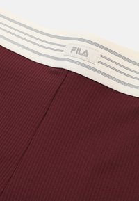 Mörkröd ribbad tyg med ett vitt midjeband som har grå ränder. Fila-logotypen är synlig på midjebandet.