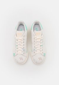 Baskets en toile blanches avec des accents bleu clair, présentant des lacets, des logos et des graphiques en vert et marron sur les côtés et les orteils.