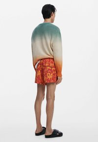 Sweter dziany w ombre odcieniu od turkusowego do pomarańczowego, połączony z czerwonymi szortami z wyrazistym żółtym wzorem graficznym oraz czarnymi sandałami.