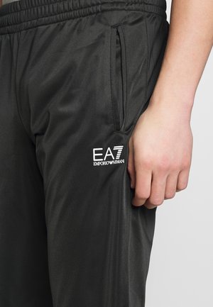 Schwarze Sporthose mit Reißverschlusstasche an der Seite und weißem EA7 Emporio Armani Logo, nah an einer entspannten Hand an der Seite.