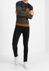 Tröja i marinblått, grått och brunt med rund hals, ribbstickade manschetter och bröstficka; matchad med svarta slim-fit jeans och vita sneakers.