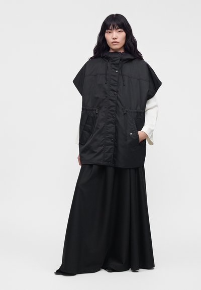Femme portant un parka noir sans manches à capuche avec de grandes poches par-dessus un haut blanc à manches longues et un pantalon noir large, debout devant un fond uni.