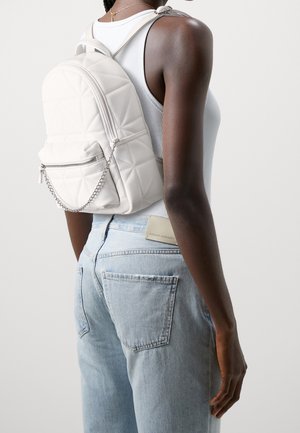 Mørkhudet person iført en hvid tanktop og lyseblå jeans, bærende på en quiltet hvid rygsæk med en lynlåslomme foran og kædedetalje.