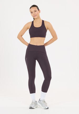 Kvinde står med hænderne bag ryggen iført mørkelilla sportstop, matchende leggings, hvide sokker og sølvfarvede løbesko.
