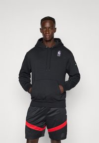Black hoodie with front pocket, drawstring hood, NHL logo patch. Worn with black shorts featuring red stripes. Soft fabric, relaxed fit.