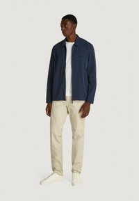 Giacca leggera blu navy con zip frontale, colletto a punta e taschino sul petto, abbinata a una t-shirt bianca e pantaloni beige, indossata con sneakers bianche.