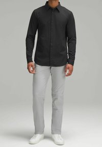 Camicia nera a maniche lunghe con bottoni e colletto, abbinata a pantaloni grigio chiaro e sneakers bianche, con una texture liscia e un design aderente.