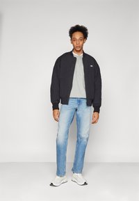 Svart bomberjakke med ribbestrikkede mansjetter, grå genser under, lyseblå jeans og hvite sneakers med mørke detaljer.