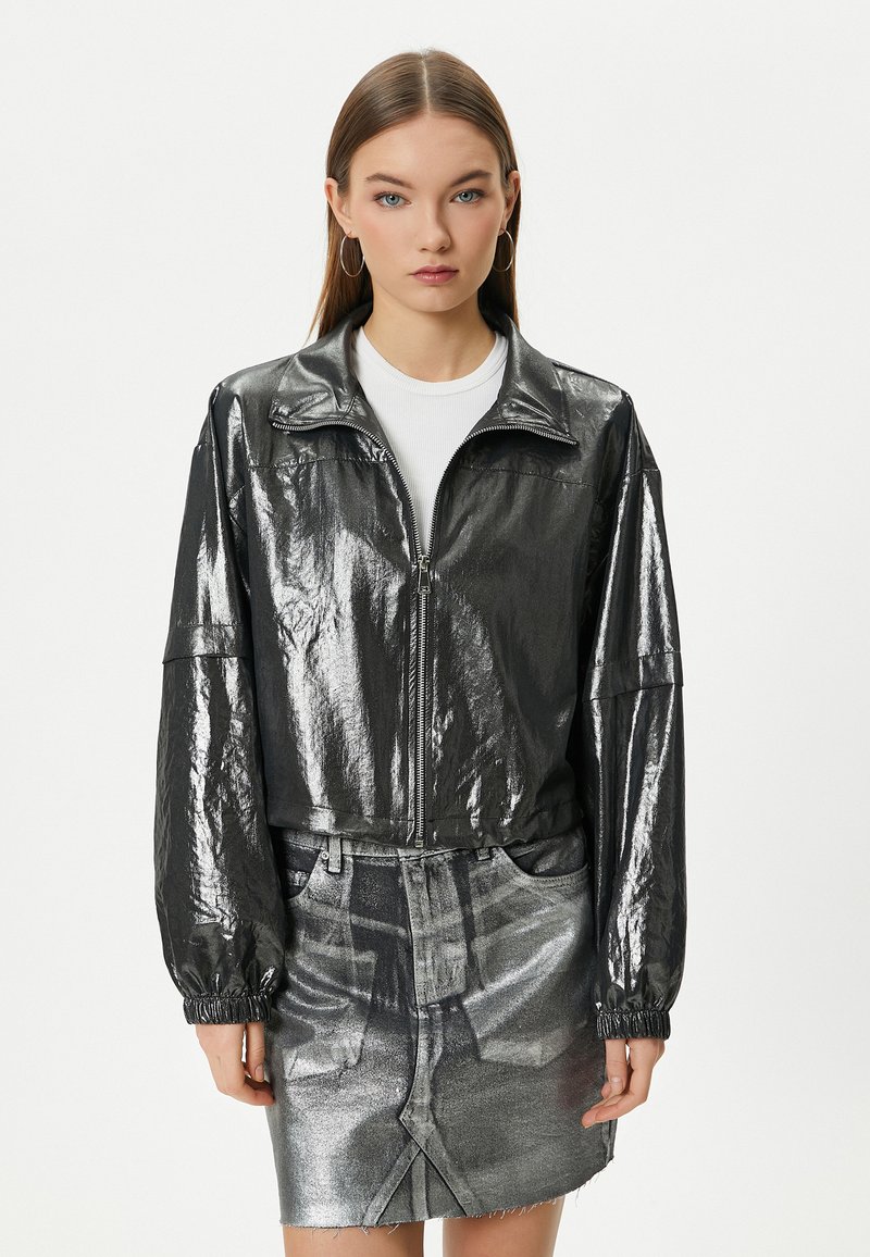 Giacca nera metallizzata con zip e colletto rialzato, polsini elasticizzati e vestibilità cropped, abbinata a una gonna di denim argentata coordinata.