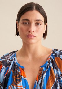 Blusa colorata con un motivo floreale nelle tonalità di blu, arancione e marrone. Il tessuto è leggero, con scollatura arricciata e maniche corte.