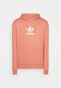 Ljus persikofärgad huvtröja tillverkad av mjukt tyg, med en känguruficka och en vit Adidas-logotyp på bröstet.