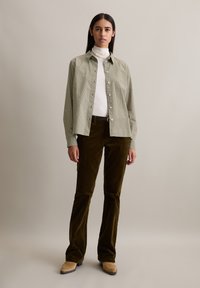 Groen en wit geruite button-up shirt, witte coltrui, olijfgroene ribfluwelen broek en beige enkellaarsjes, staande tegen een neutrale achtergrond.