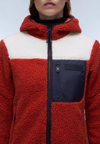 Red and cream fleece jacket with a zippered navy pocket. Features a hood, textured fabric, and color block design.