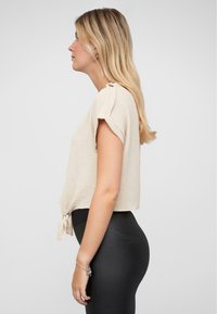 Top corto beige con maniche corte, tessuto testurizzato e dettaglio con nodo laterale. Indossato con leggings neri. Vista di profilo laterale.