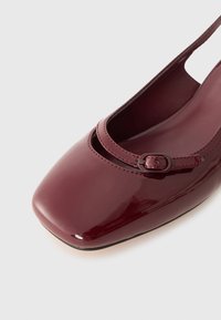Ballerine en cuir verni bourgogne avec un bout carré arrondi, dotée d'une fine sangle et d'un détail de boucle ovale. Texture lisse.
