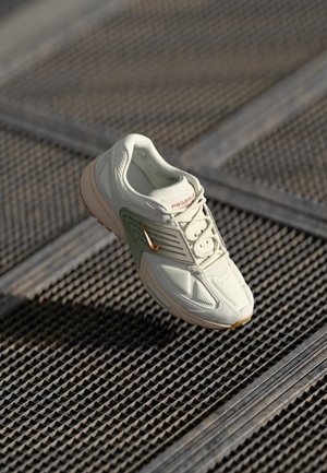 Jasnobeżowy i zielony but do biegania Nike Pegasus z miedzianym znakiem Swoosh, leżący na metalowej siatce z liniami diagonalnymi.