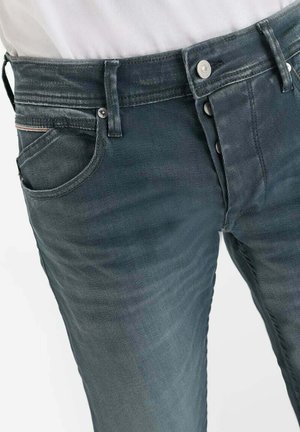 Close-up van donkergrijze spijkerbroek met knoopsluiting, voorzak en riemlussen, gedragen met een ingestopte witte blouse.