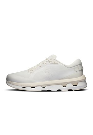White low-top athletic sneaker with textured mesh upper, beige midsole, unique cutout details in the sole, and lace-up closure, side view.