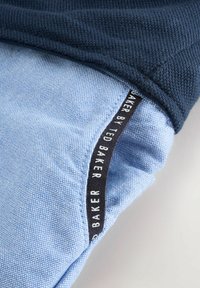 Jūras zila trikotāžas audums pārklājas ar teksturētu gaiši zilu materiālu, uz kura atrodas melna etiķete ar uzrakstu "BAKER BY TED BAKER". Redzama šuvju detaļas un krāsu kontrasts.