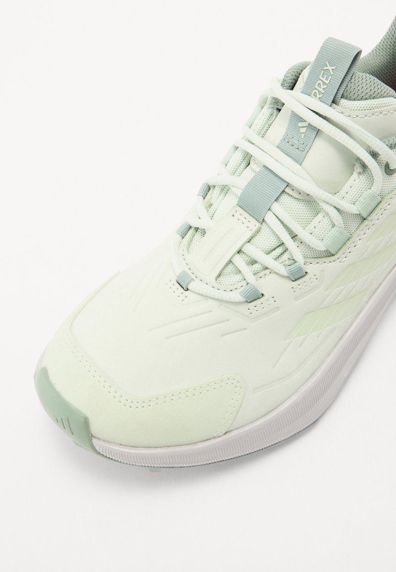 Zapatilla deportiva de color verde claro con parte superior texturizada, cordones blancos y una sutil marca. Presenta una punta moldeada y una suela blanca estriada.