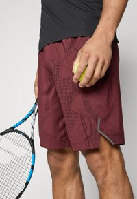 Pantalones cortos deportivos granates con un patrón geométrico, con un acento gris contrastante en el dobladillo. Sosteniendo una pelota de tenis verde y una raqueta.