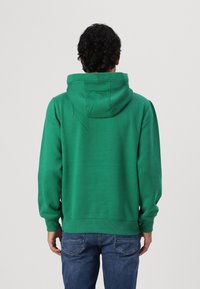 Grüne Kapuzenjacke aus weichem Stoff, mit Kordelzug-Kapuze, gerippten Bündchen und Saum, von hinten gezeigt. Getragen mit blauen Jeans.