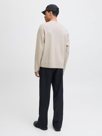 Man som bär beige tröja, svarta vida byxor, svarta skor och svart keps, stående med ryggen mot en enfärgad bakgrund.