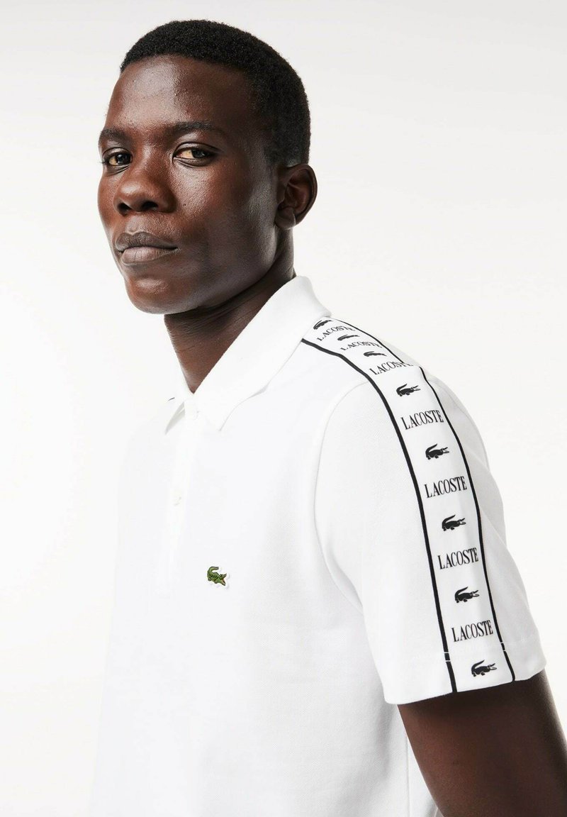 Polo shirt blanc à manches courtes, avec un logo crocodile vert et des détails en bande noire arborant le logo Lacoste répété sur les épaules.