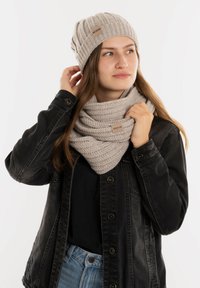 Lichtgrijze gebreide beanie en infinity-sjaal, met een gemêleerd patroon en leren labels, gedragen met een zwarte spijkerjacket.