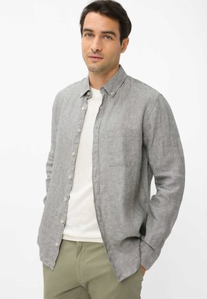 Mann trägt graues Hemd mit Knöpfen über weißem T-Shirt und khakifarbene Hose, steht mit Händen in den Taschen vor schlichten Hintergrund.