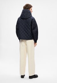 Marineblau gesteppte Jacke mit Kapuze, ausgestattet mit einem Rautenmuster und elastischen Bündchen. Kombiniert mit cremefarbenen weitem Hosen und schwarzen Schuhen.