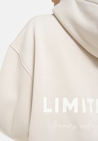 Beige Kapuzenpullover mit großem weißen Text "LIMITED" und kleinerer kursiver Schrift "dreams endless" auf dem Rücken, getragen von einer Person.