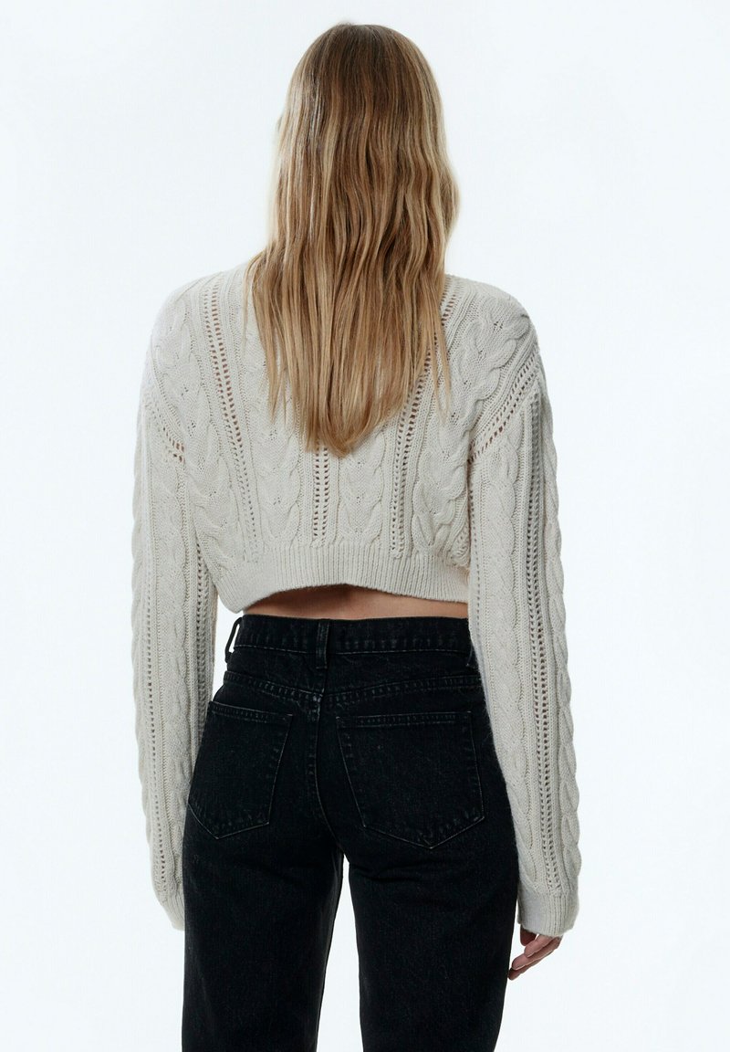 Pull en maille torsadée crème, court, avec des manches longues, présentant des motifs texturés. Associé à un jean noir taille haute, vue de dos.