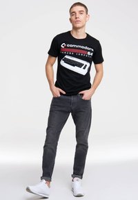 Black cotton t-shirt featuring a retro Commodore 64 gaming computer graphic. Worn with gray jeans and white sneakers.