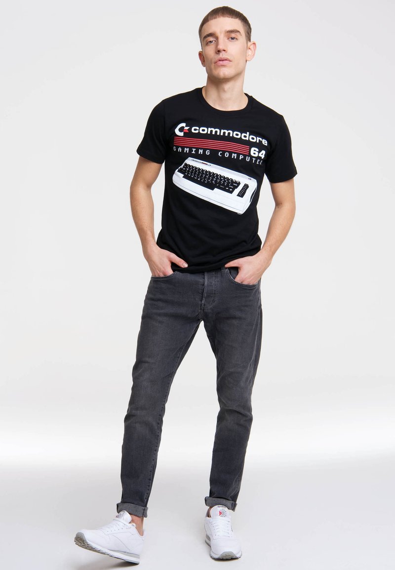 Black cotton t-shirt featuring a retro Commodore 64 gaming computer graphic. Worn with gray jeans and white sneakers.
