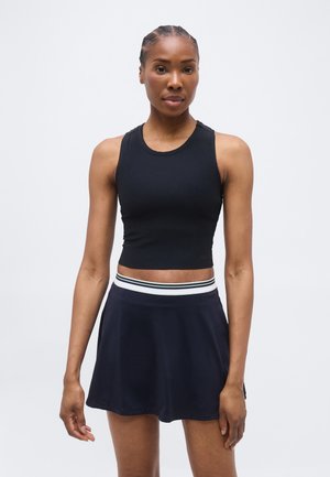 Γυναίκα που φοράει μαύρο αμάνικο κοντό τοπ και ναυτικό μπλε φούστα με λευκή ρίγα στη μέση, στέκεται μπροστά σε απλό φωτεινό φόντο.