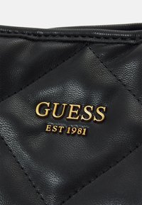 Borsa a tracolla in pelle trapuntata nera con logo oro "GUESS" e testo "EST 1981". Presenta una texture liscia e dettagli di cucitura puliti.