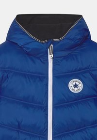 Blå puffjacka med huva, med en vit dragkedja och en cirkulär Converse-logotyp på vänster bröst. Blankt, isolerat material.