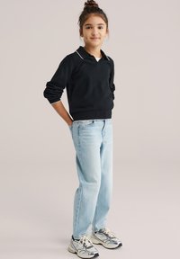 Jeune fille debout, mains derrière le dos, portant un haut noir à manches longues, un jean bleu clair et des baskets argentées, sur un fond uni.