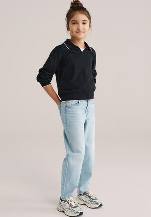 Giovane ragazza in piedi con le mani dietro la schiena, indossa una maglietta nera a maniche lunghe, jeans azzurri chiari e sneakers argentate su uno sfondo semplice.