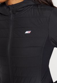 Närbild av en kvinna i en svart vadderad jacka med dragkedja fram och en liten rektangulär logotyp i rött, vitt och blått på bröstet.