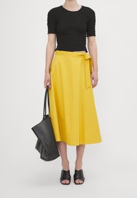 Top corto nero e texturizzato abbinato a una gonna giallo brillante fino al ginocchio con una cintura a fiocco. La modella tiene una borsa tote nera e indossa sandali neri.