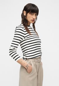 Ριγέ μακρυμάνικο πουκάμισο σε λευκό και μαύρο, με στρογγυλή λαιμόκοψη και στενή εφαρμογή· φορεμένο με μπεζ παντελόνια ψηλόμεσα και χέρια στις τσέπες.